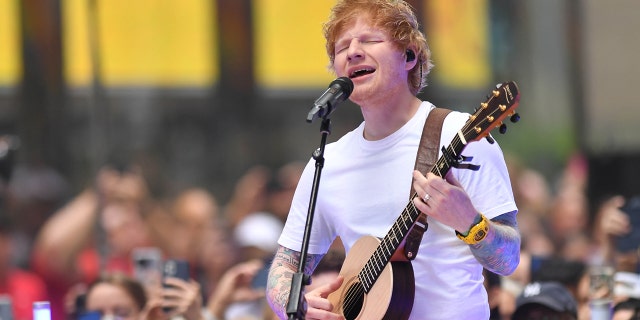 Ed Sheeran sings into the microphone while playing the guitar and closing his eyes in NYC