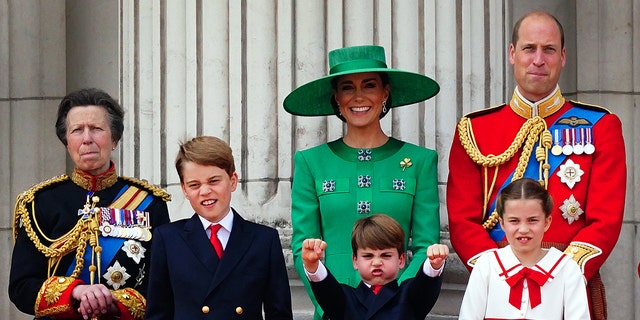 The Princess Royal stands beside Prince George, who display his own funny face, next to Prince Louis outstretching his arms and showing his fists, with Kate Middleton laughing behind her son, and Prince William trying to hold in a laugh, in front of Princess Charlotte