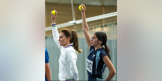 Kate Middleton holds a ball up in the air as if she were to feed it to the player on the baseline next to another young girl