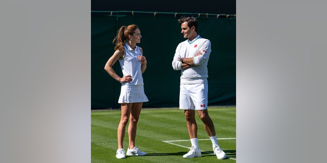 Kate Middleton with her hands on her hips in white talks with Roger Federer on the court of Wimbledon