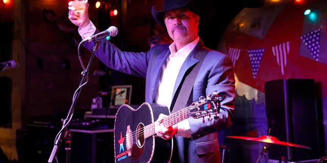 John Rich raises a glass on stage with a guitar swung across his chest in Nashville