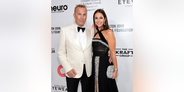 Kevin Costner soft smiles in a white tuxedo with his wife Christine in a black criss-cross gown at the Elton John AIDS Foundation party