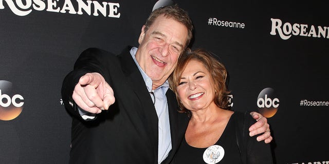 John Goodman points at the camera and smiles next to Roseanne Barr in a black dress with a white flower on it