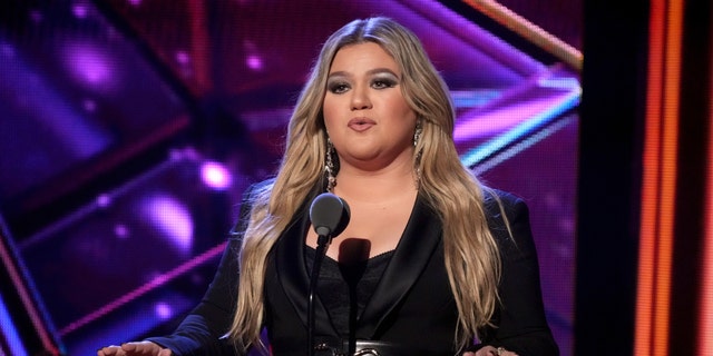 Kelly Clarkson in a black outfit behind a microphone at the iHeartRadio Music Awards