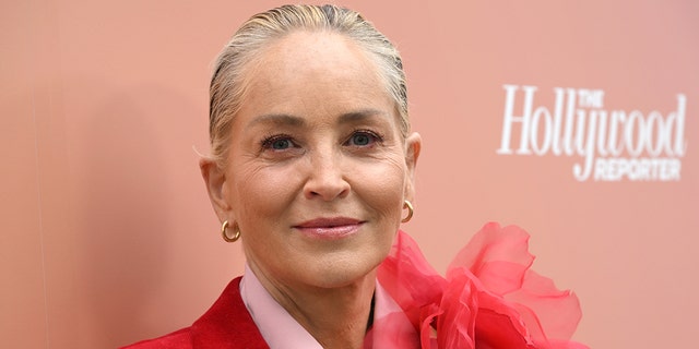 Sharon Stone soft smiles on the red carpet at The Hollywood Reporter Raising Our Voices DEIA Luncheon wearing a red suit with a large flower on the shoulder