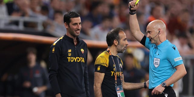 Anthony Taylor shows a yellow card to a Roma coach