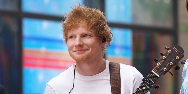 Ed Sheeran smiles wearing a white shirt in NYC with his guitar strapped over his arm and earpiece hanging on his shirt