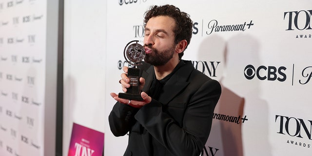 Brandon Uranowitz in a black jacket kisses his Tony Award