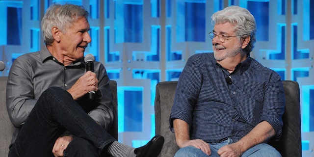 Harrison Ford sits on a couch holding a microphone looking at George Lucas who is laughing for a Star Wars event