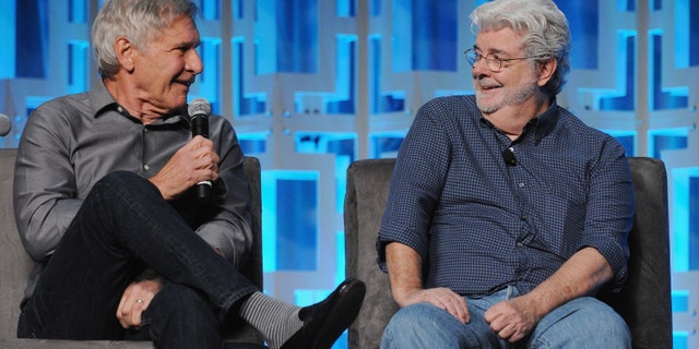Harrison Ford sits on a couch holding a microphone looking at George Lucas who is laughing for a Star Wars event
