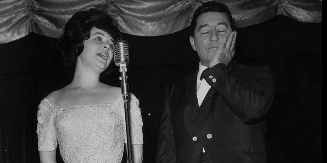 Louis Prima closing his eyes and holding his hand at Gia Maione wearing a white dress sings to the mic