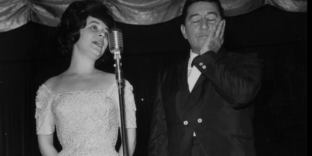 Louis Prima closing his eyes and holding his hand at Gia Maione wearing a white dress sings to the mic