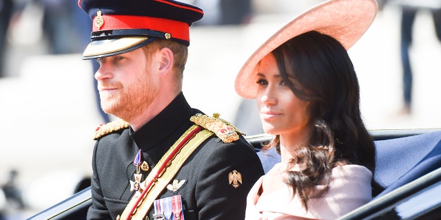 A close-up of Meghan Markle wearing a pink dress and matching hat sitting next to Prince Harry in his military uniform