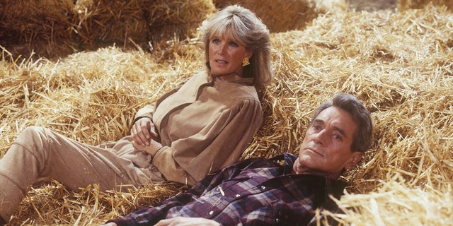 Linda Evans wearing a tan dress laying on hay with Rock Hudson