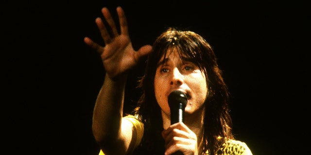 Steve Perry sings into the microphone wearing a yellow shirt, reaching out into the air with his hand