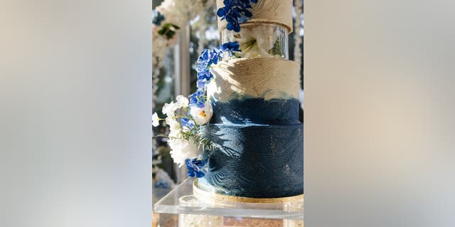 Wedding cake with a base tier that has swirling blue icing that eventually switches to white is on display. The cake has decorative gold flecks and blue and white flowers.