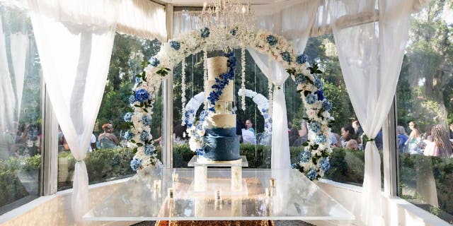 A five-tier wedding cake with a blue base that fades into white. There's an acrylic cake separator in the middle. The cake is set up inside an indoor ballroom and sits in front of a flower arch.