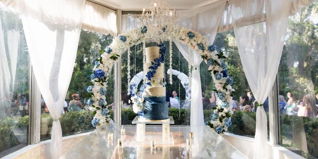 A five-tier wedding cake with a blue base that fades into white. There's an acrylic cake separator in the middle. The cake is set up inside an indoor ballroom and sits in front of a flower arch.