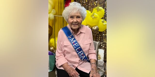Dillard's retiree sitting in a chair in front of yellow balloons and a gold curtain she is wearing a pink shirt and a sash that says melba