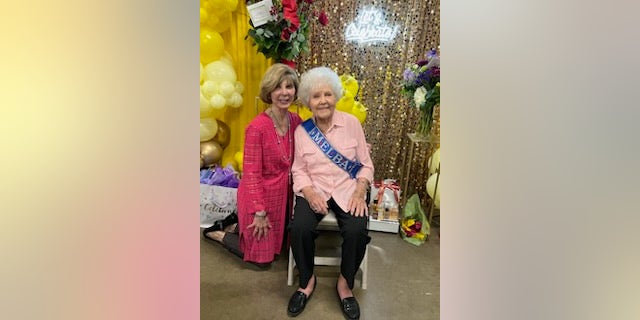 Melba wearing a pink shirt is sitting next to drew dillar who is wearing a pink cardigan, they are both in front of a gold curtain and yellow baloons.