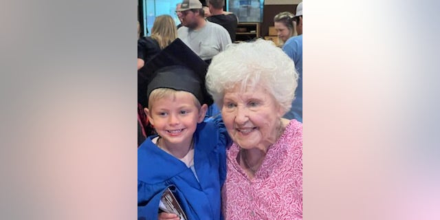 Young boy on the left held up by his grandmother on the right, the boy is wearing a graduation gown