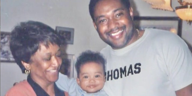 Minnie Smith is holding a smiling baby next to her smiling grandson wearing a grey sweatshirt