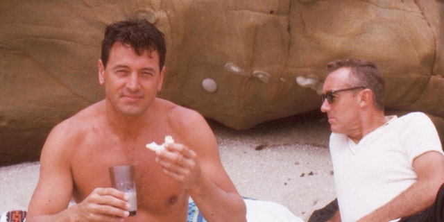 Rock Hudson shirtless on the beach next to a male friend wearing a white shirt and sunglasses