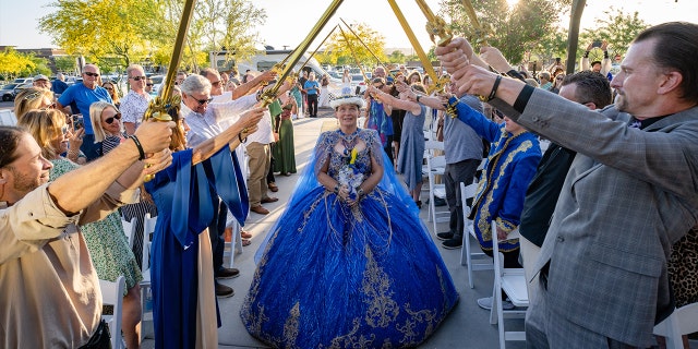 Jamie West walks down the aisle in a blue and gold wedding gown and crown while guests hold up decorative swords.