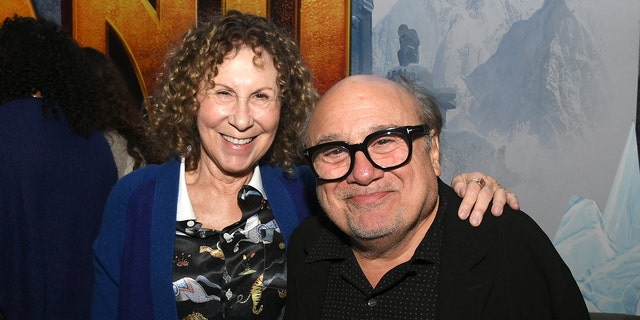 Rhea Perlman in a blue cardigan and printed shirt puts her arm around Danny DeVito in a black shirt and black glasses