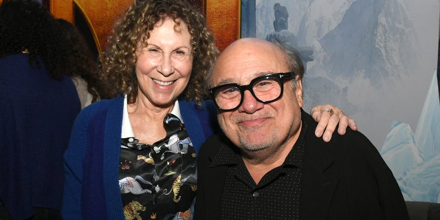 Rhea Perlman in a blue cardigan and printed shirt puts her arm around Danny DeVito in a black shirt and black glasses