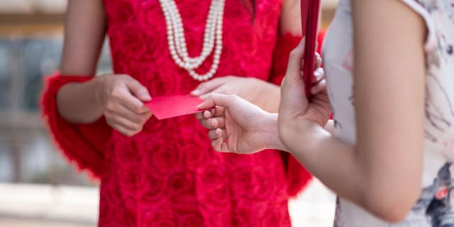 Woman in red dress and pearl necklace hands a red envelope to another woman in a floral white dress.