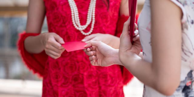 Woman in red dress and pearl necklace hands a red envelope to another woman in a floral white dress.