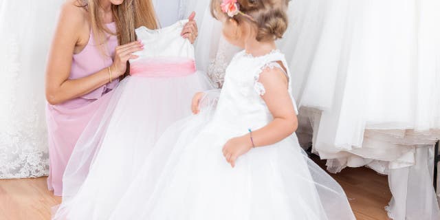 Woman holds up a flower girl dress in front of a young girl.
