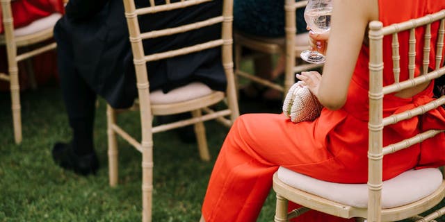 Woman wears a red jumpsuit while sitting on a lawn chair during what appears to be a wedding ceremony. Another woman a short distance can be seen wearing red as well.