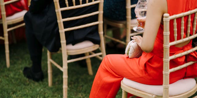Woman wears a red jumpsuit while sitting on a lawn chair during what appears to be a wedding ceremony. Another woman a short distance can be seen wearing red as well.