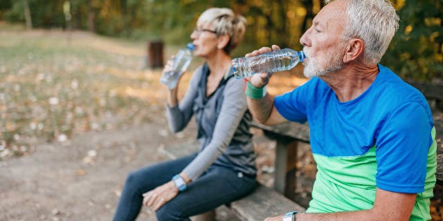 An older man and woman stop to drink water while sitting on a park bench.