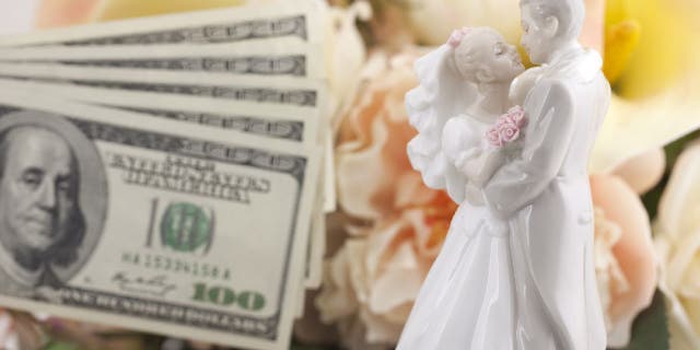 Hundred dollar bills next to bride and groom cake topper and bouquet.