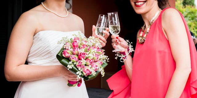 A bride wears a white dress and holds a bouquet while she toasts champagne with a wedding guest who's wearing a red dress. 