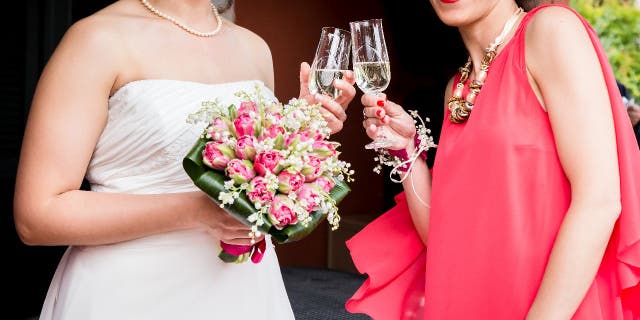 A bride wears a white dress and holds a bouquet while she toasts champagne with a wedding guest who's wearing a red dress.