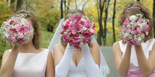 Bride poses with two bridesmaids in pink dresses while each of them hold a bouquet in front of their faces.