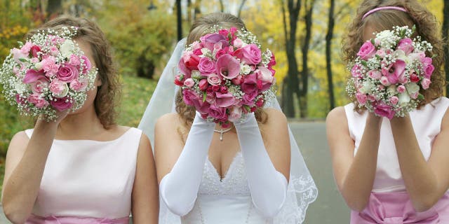 Bride poses with two bridesmaids in pink dresses while each of them hold a bouquet in front of their faces.
