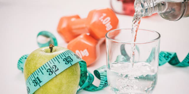 Glass of water gets filled next to green apple, dumbbells and measuring tape.