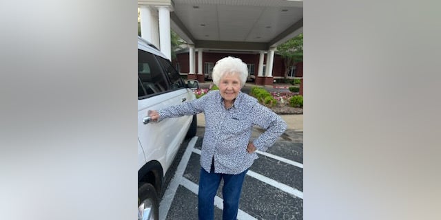 Older woman standing next to a car in front of a building grabbing a car door handle