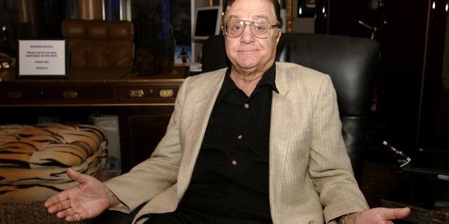 Pat Cooper sits on a leather chair while wearing a khaki blazer