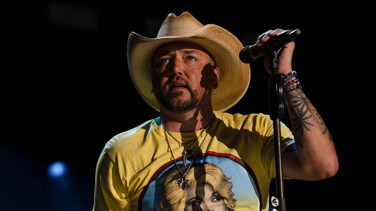 Jason Aldean in a yellow shirt with Dolly Parton on it and a tan cowboy hat holds the microphone stand