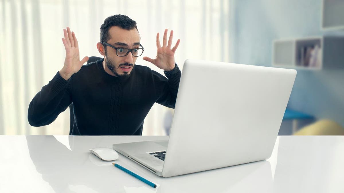 Man looking shocked, throwing hands in air while looking at laptop