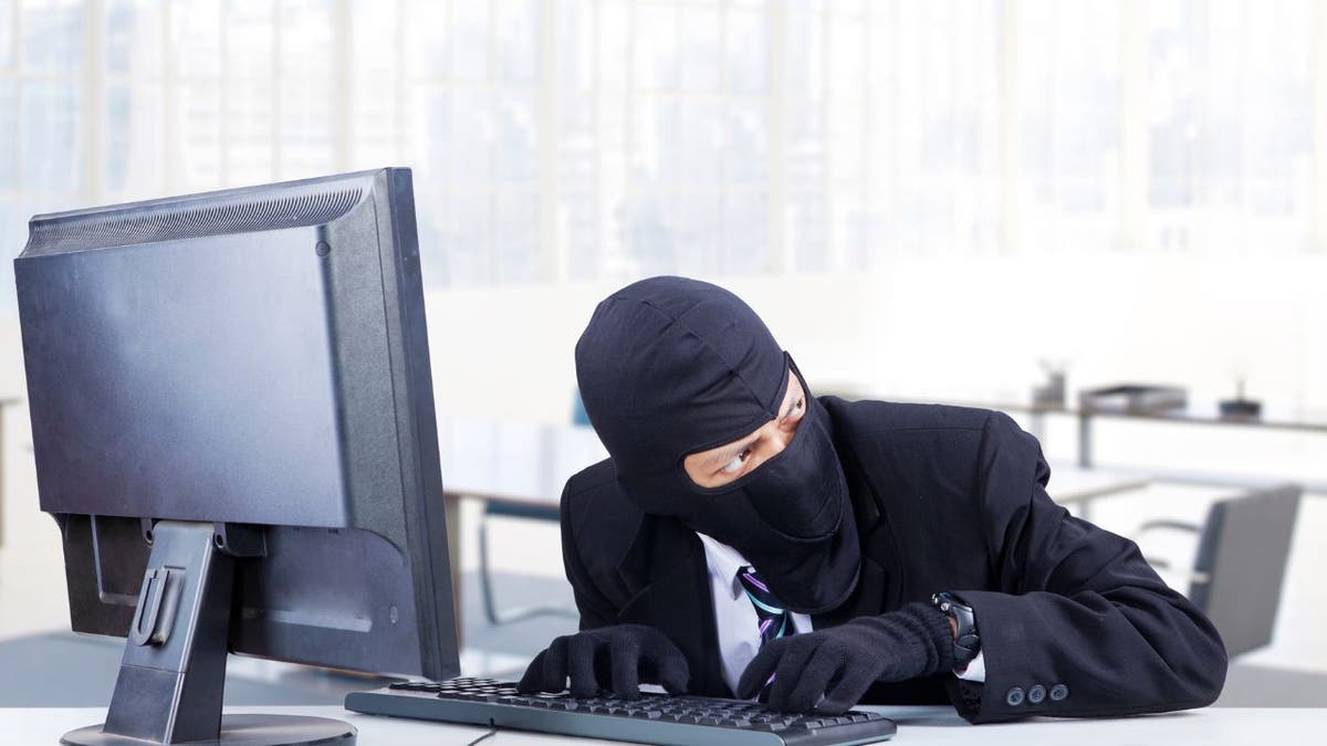 Hacker in black suit, gloves, and mask, typing on computer, looking to the right