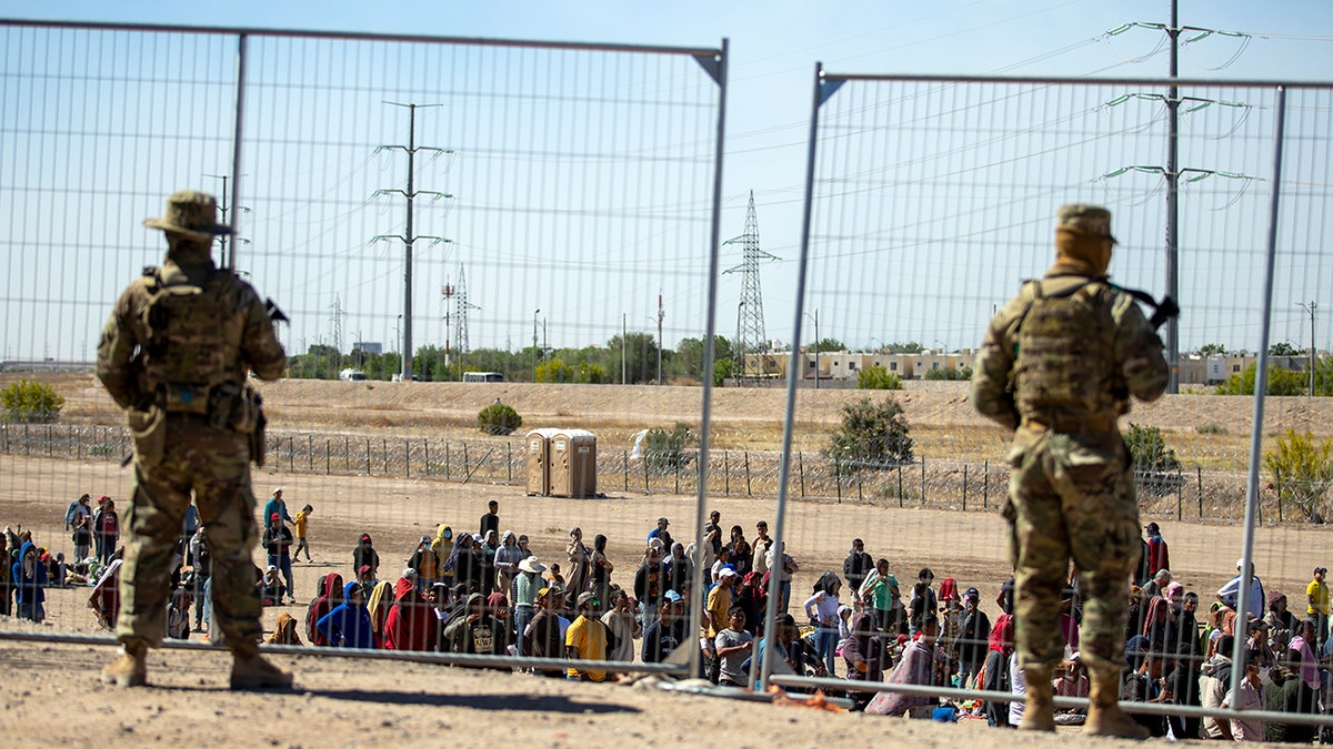 Migrants wait in line adjacent to the border fence 