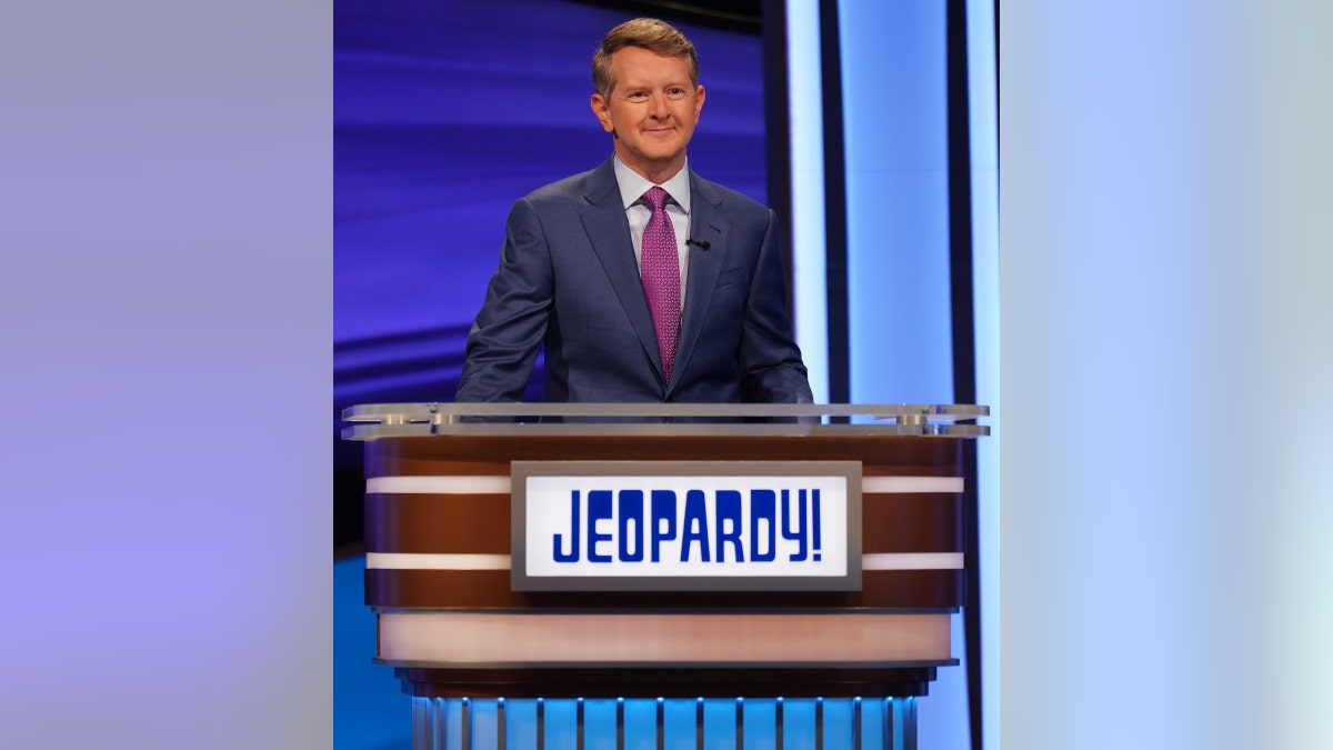 Ken Jennings in a blue suit with a purple tie behind the Jeopardy! podium