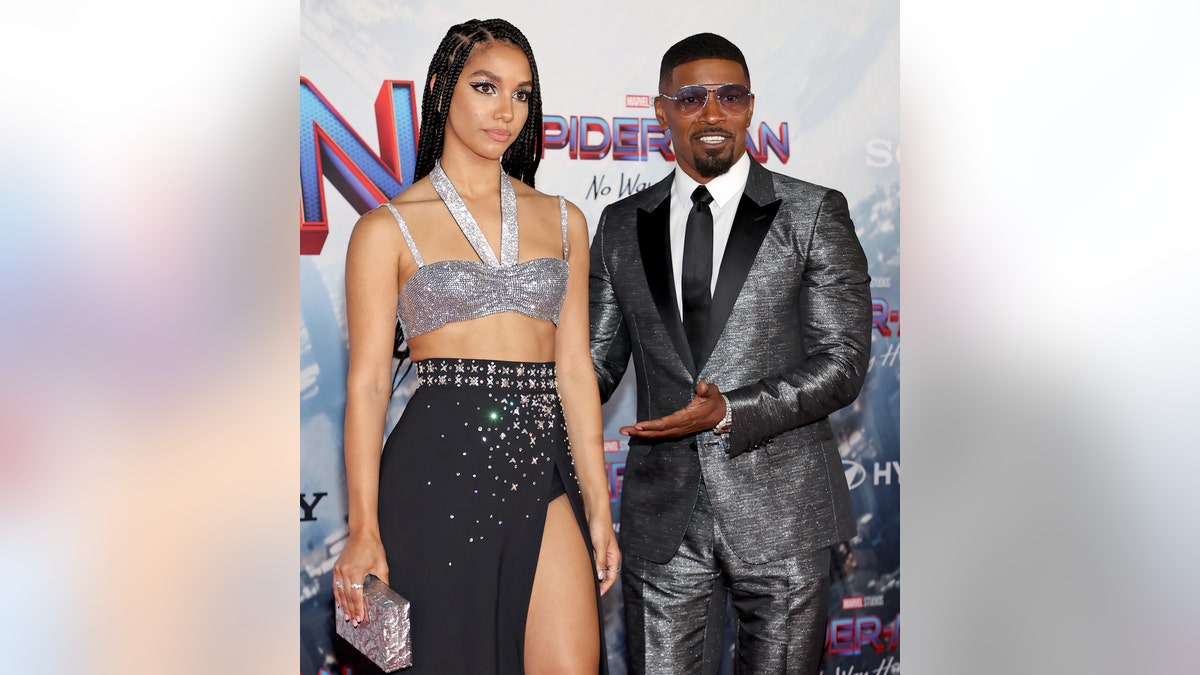 Corinne Foxx in a two piece outfit stands next to Jamie Foxx who emphasizes here